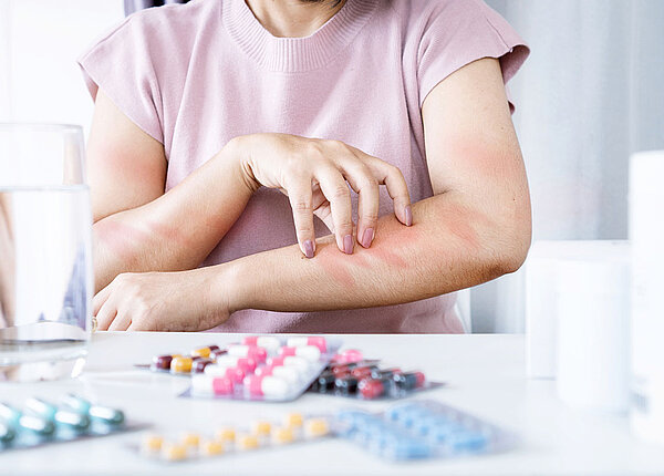 Eine Frau sitzt am Tisch. Vor ihr liegen zahlreiche Tabletten- oder Kapselblister. Sie kratzt sich am Arm.