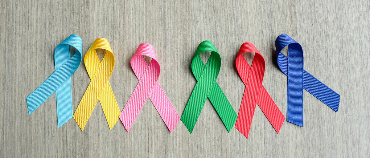 Auf einem Holzuntergrund liegen nebeneinander sechs Krebs-Awareness-Schleifen in verschiedenen Farben: hellblau, gelb, rosa, grün, rot und dunkelblau.