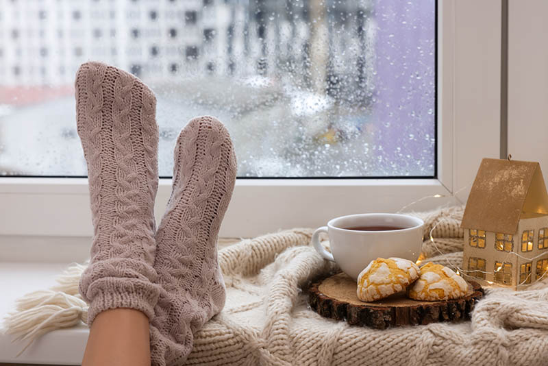 Eine Frau hat die Füße in Stricksocken auf die Fensterbank gelegt. Daneben stehen Tee, Kekse und ein beleuchtetes Deko-Haus. Es ist Tag, aber trotzdem nicht richtig hell. Es schneit.