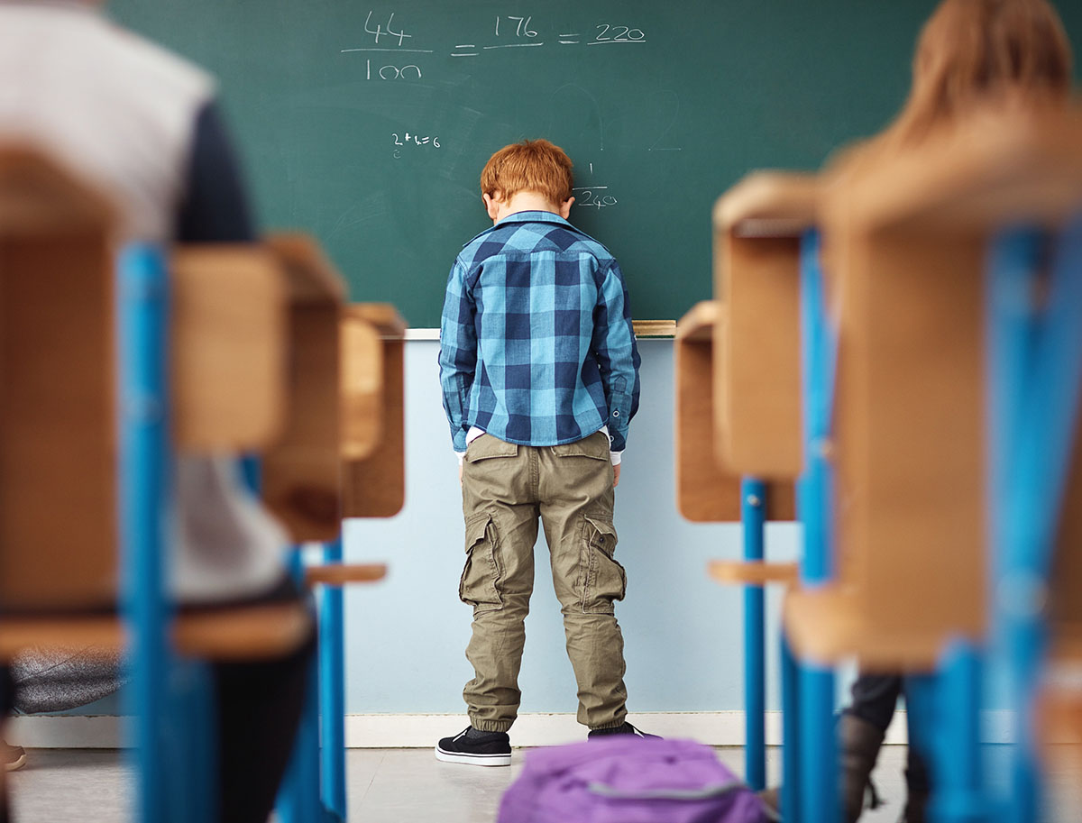 Ein Junge im Grundschulalter steht vor der der Schultafel, lässt den Kopf gegen die Tafel sinken und hat die Hände in den Hosentaschen.