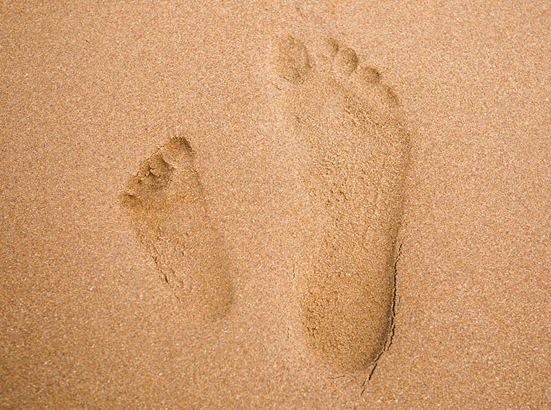 Ein Kinder- und ein Erwachsenenfußabdruck im Sand. Der Kinderabdruck ist sehr flächig. Beim Erwachsenenabdruck ist die Innenseite gewölbt.