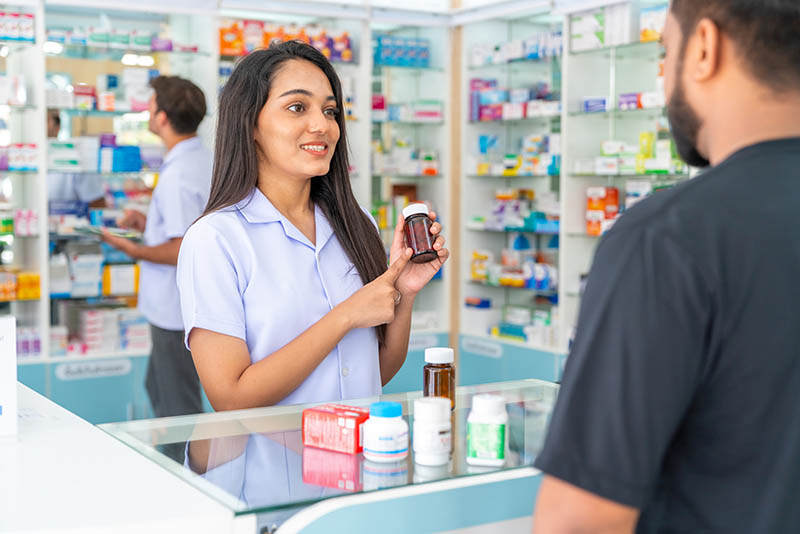 Beratung in der Apotheke: Auf dem HV-Tisch stehen 5 verschiedene Packungen mit Nahrungsergänzungsmitteln. Eine PTA oder Apothekerin hält eine sechste Packung in der Hand und erklärt ihrem Kunden etwas.