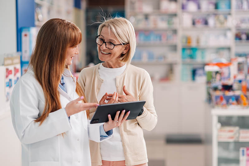 Eine PTA oder Apothekerin berät eine Kundin etwas abseits vom HV-Tisch mit Hilfe eines Tablets. Beide sehen sehr zufrieden aus.