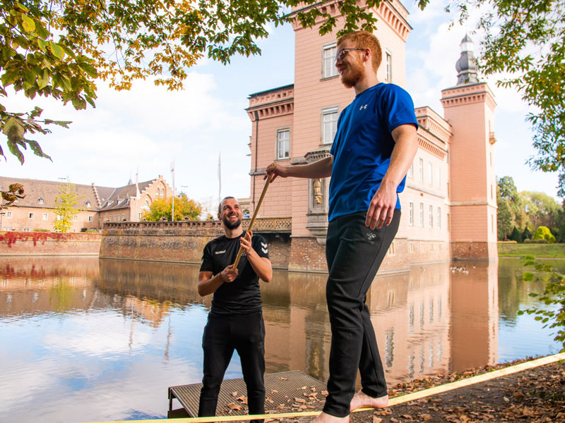 Ein Mann balanciert auf einer Slackline, ein weiterer Mann unterstützt ihn. Im Hintergrund ist Schloss Gracht zu sehen.