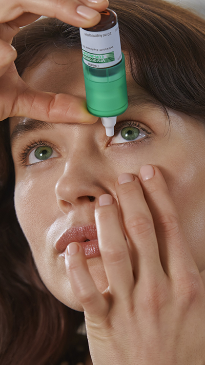 Eine Frau tropft sich Visiodoron Euphrasia® Augentropfen aus einem grün-weißen Mehrdosenbehältnis ins Auge.