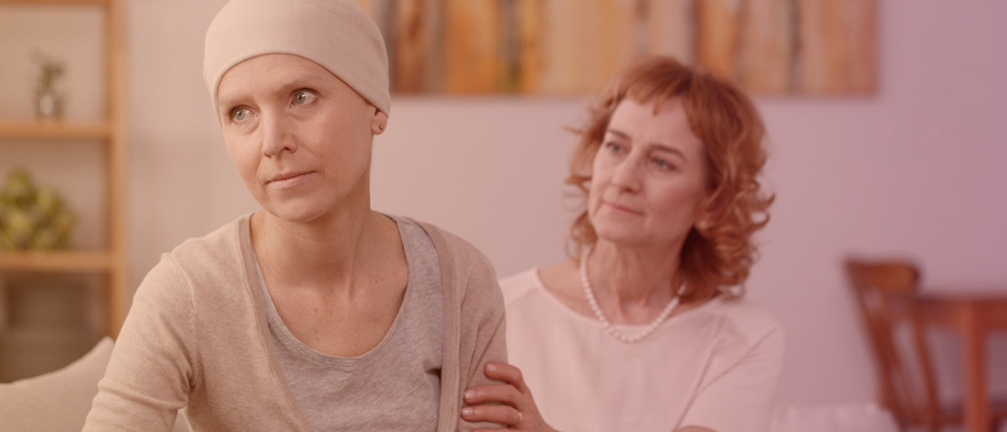 Krebsbehandlung und Nebenwirkungen von Krebsmedikamenten: Allgemeine Beratungstipps rund um die Therapie Eine Krebspatientin mit Mütze sitzt auf dem Sofa und schaut in die Ferne. Eine weitere Frau sitzt hinter ihr, schaut sie an und hält ihren Arm.