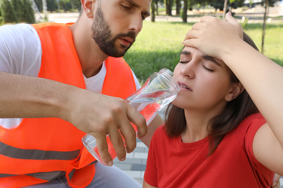 Erste Hilfe bei Hitzschlag und Sonnenstich: Symptome erkennen und Maßnahmen ergreifen Ein Sanitäter reicht einer Frau eine Flasche Wasser. Sie sitzt auf dem Boden, schaut gequält und hält sich die Hand an den Kopf.