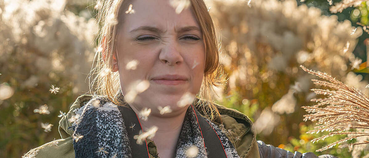 Eine Frau steht im Freien und Pappelpollen fliegen an ihrem Gesicht vorbei. Sie verkneift das Gesicht.