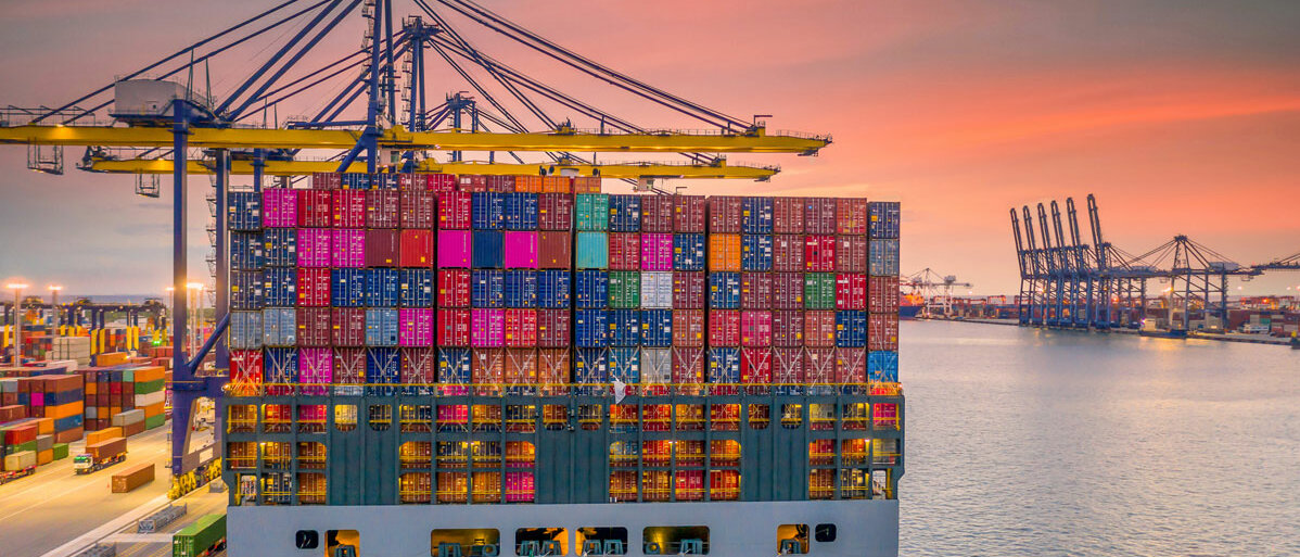 Ein Containerschiff, beladen mit vielen bunten Containern, liegt in einem Hafen mit mehreren Kränen. Der Himmel ist rosa, weil die Sonne gerade auf- oder untergeht.