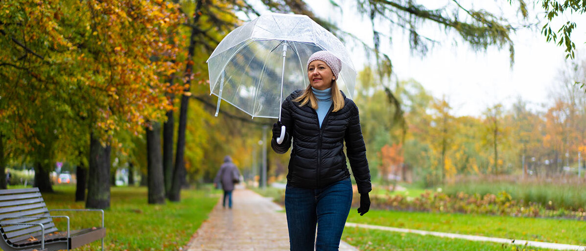Frau mit Regenschirm läuft bei wechselhaftem Wetter durch einen Park.
