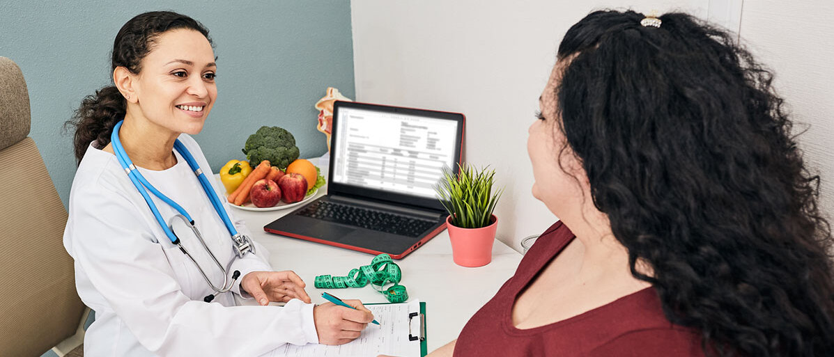 Eine Frau im weißen Kittel spricht mit einer dicken Patientin. Auf dem Tisch steht eine Schale mit Obst und Gemüse.