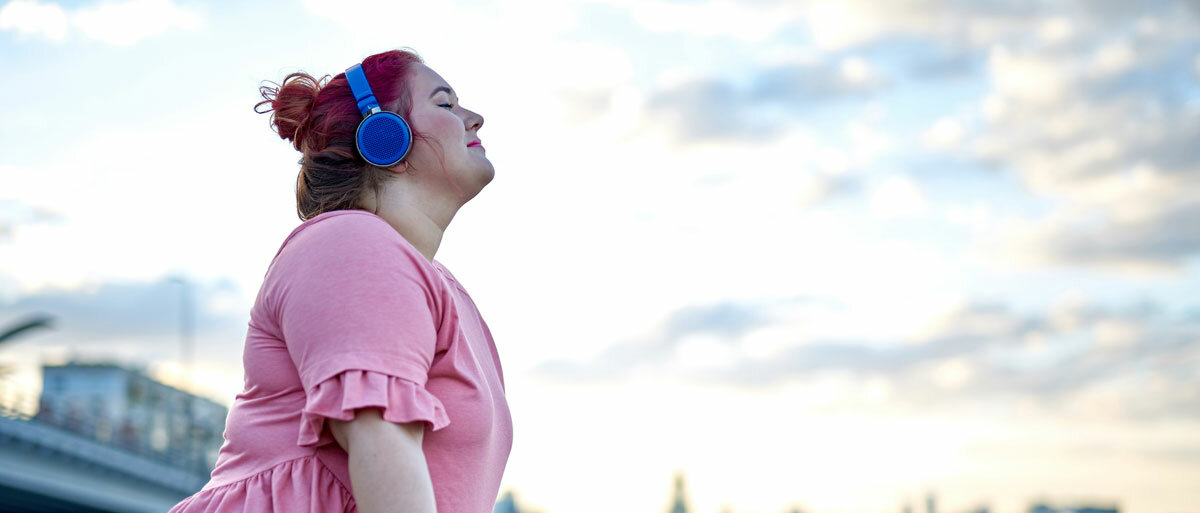 Eine Frau lehnt mit geschlossenen Augen an einem Brückengeländer und genießt die Musik, die sie über ihre Kopfhörer hört.