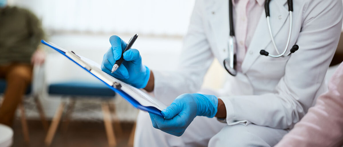 Ein Arzt in Kittel und Schutzausrüstung sitzt neben einem Patienten oder einer Patientin und notiert etwas auf einem Klemmbrett.