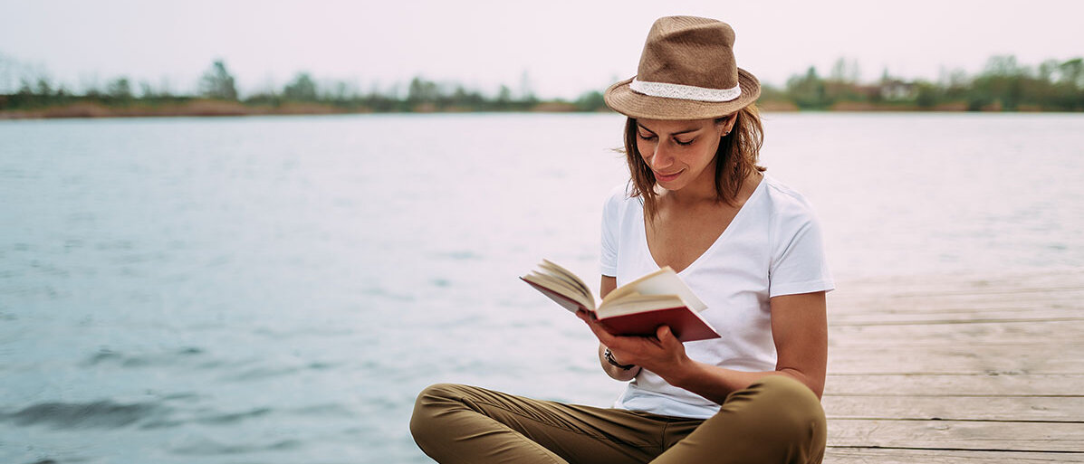 Frau sitzt auf einem Steg am Wasser und liest ein Buch. Sie hat einen Hut auf