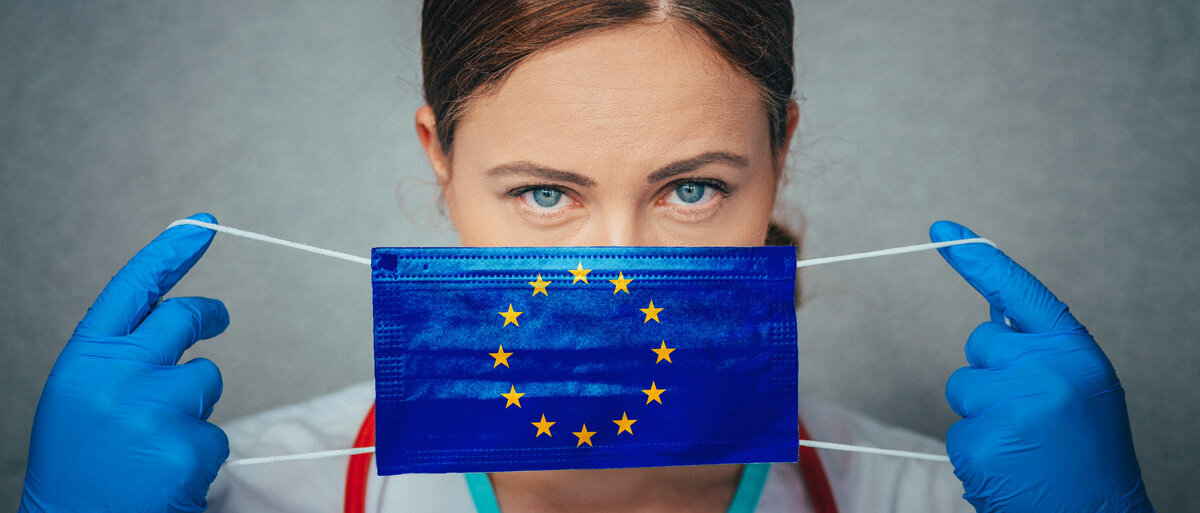Eine junge Ärztin oder Pflegerin mit blauen Augen und blauen Handschuhen zieht eine chirurgische Maske auf, auf der die EU-Flagge abgebildet ist.