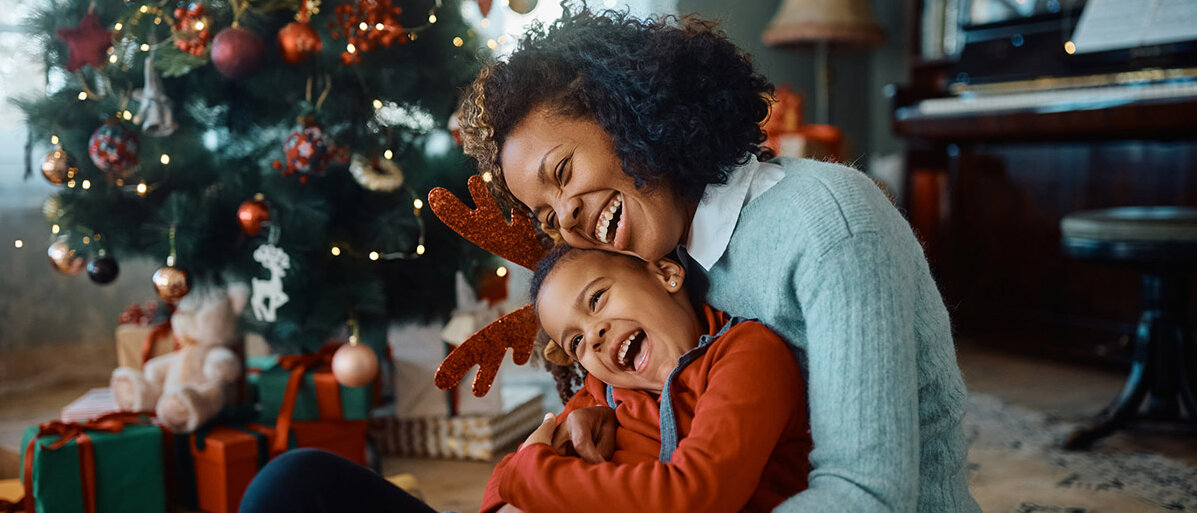 Eine Mutter und ihre Tochter sitzen neben dem Weihnachtsbaum, umarmen sich und lachen.