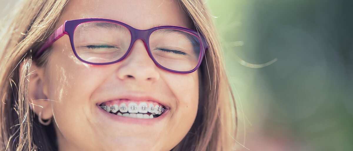 Ein Mädchen trägt eine auffällige violette Brille und eine Zahnspange. Sie lacht mit geschlossenen Augen.