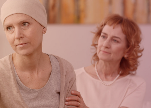 Eine Krebspatientin mit Mütze sitzt auf dem Sofa und schaut in die Ferne. Eine weitere Frau sitzt hinter ihr, schaut sie an und hält ihren Arm.