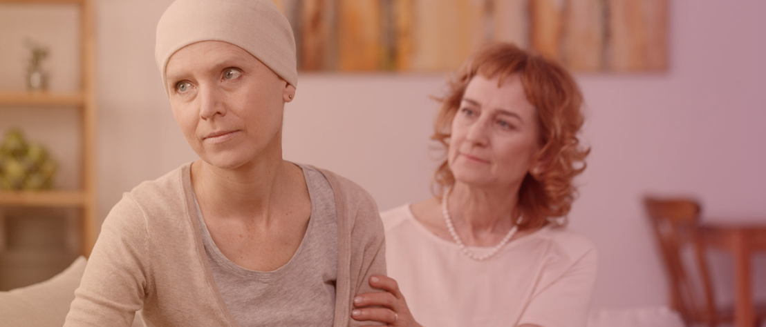 Eine Krebspatientin mit Mütze sitzt auf dem Sofa und schaut in die Ferne. Eine weitere Frau sitzt hinter ihr, schaut sie an und hält ihren Arm.