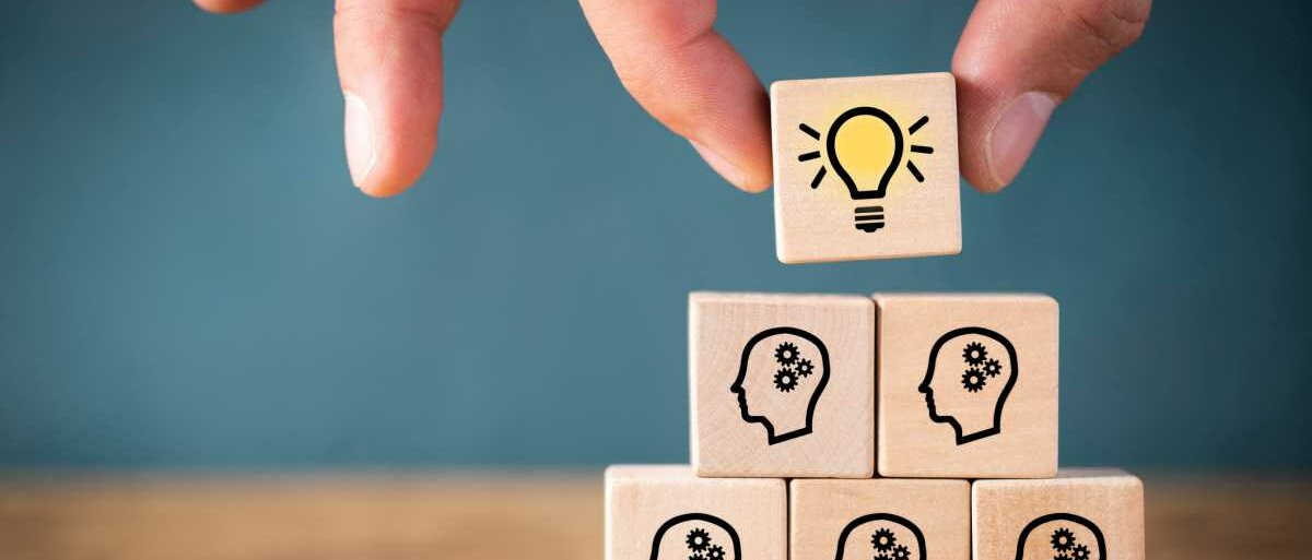 Fortbildung Mehrere Holzwürfel bilden eine Pyramide, auf ihnen ist jeweils ein Kopf mit Zahnrad als Gehirn stilisiert. Als Spitze der Pyramide wird gerade ein Würfel mit dem Icon einer leuchtenden Glühbirne aufgesetzt.