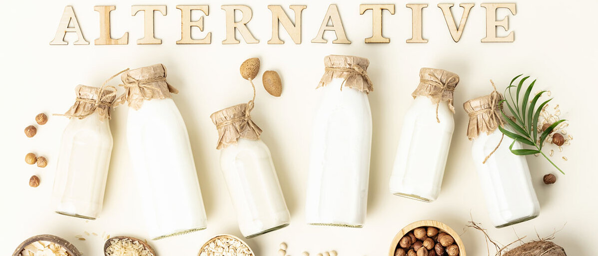Pflanzliche Milch in Flaschen und Zutaten auf hellem Hintergrund mit Holzbuchstaben „Alternative“.