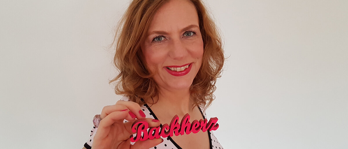 Kirsten Metternich von Wolff lächelt und hält einen roten Backherz-Schriftzug aus Holz in der Hand.
