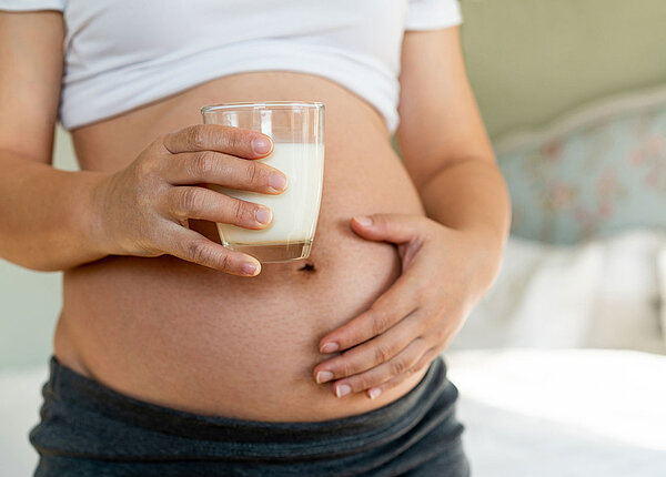 Schwangeren Bauch. Frau haelt in einer Hand ein Glas Milch und die andere Hand liegt auf dem Bauch.
