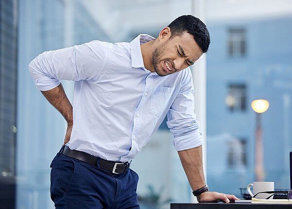 Wie Rückenschmerzen im Job und psychischer Stress zusammenhängen Geschäftsmann hat am Arbeitsplatz Rückenschmerzen und hält sich mit schmerzverzerrtem Gesicht den unteren Rücken.