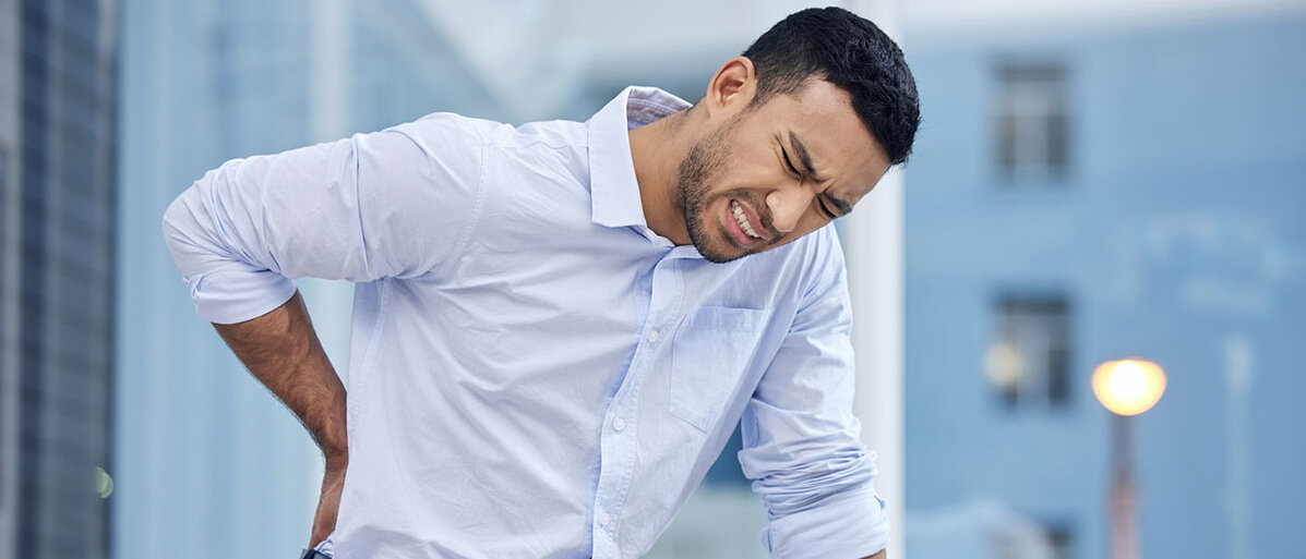 Wie Rückenschmerzen im Job und psychischer Stress zusammenhängen Geschäftsmann hat am Arbeitsplatz Rückenschmerzen und hält sich mit schmerzverzerrtem Gesicht den unteren Rücken.