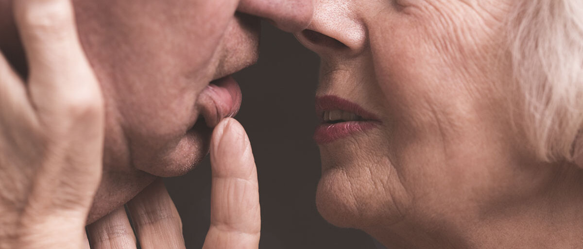 Eine Frau mit grauen Haaren und faltigem Gesicht hält das Gesicht ihres Partner nah an ihres. Einen Finger hat sie auf seine Lippen gelegt.
