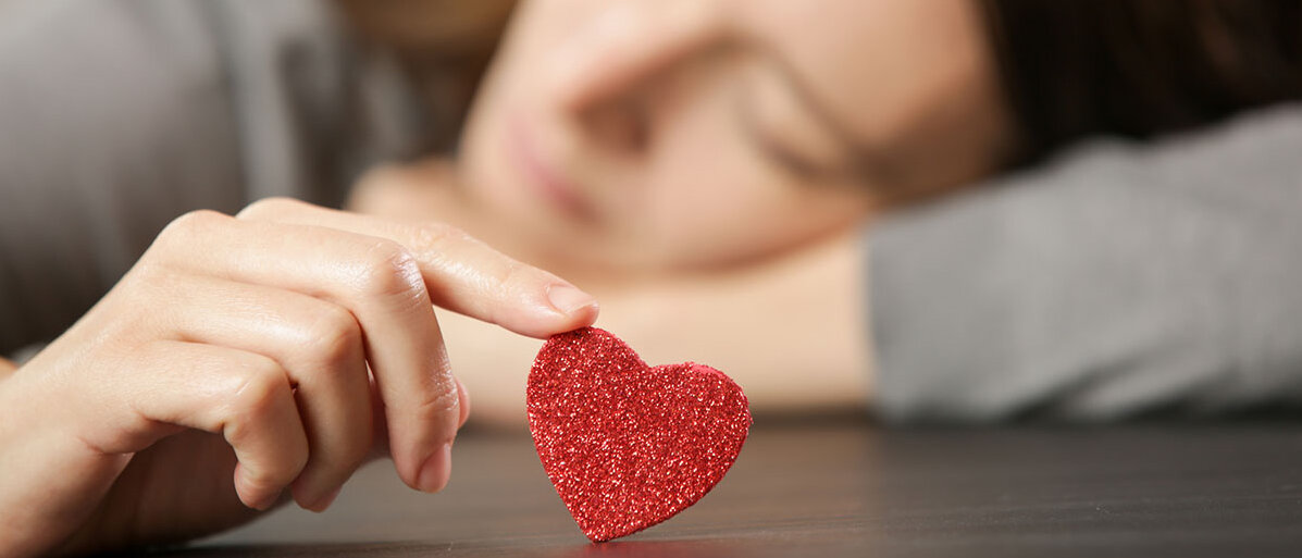 Broken-Heart-Syndrom Traurige Frau legt Kopf auf Tisch ab und spielt mit einem kleinen Herzen