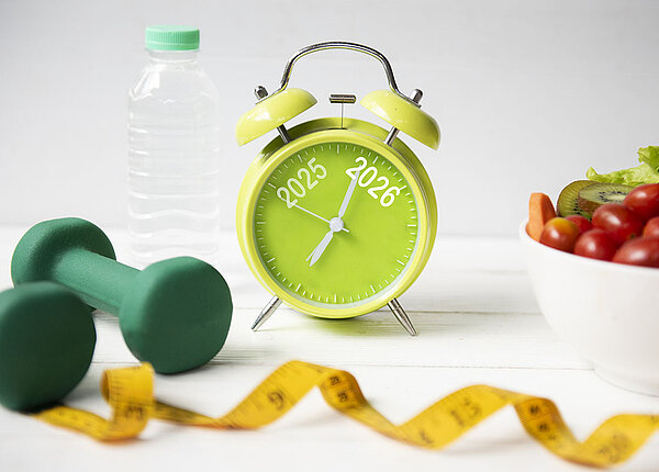 Gemüse, Obst, Hanteln und Maßband neben einer Wasserflasche und einem Wecker auf dem 2025 und 2026 steht 