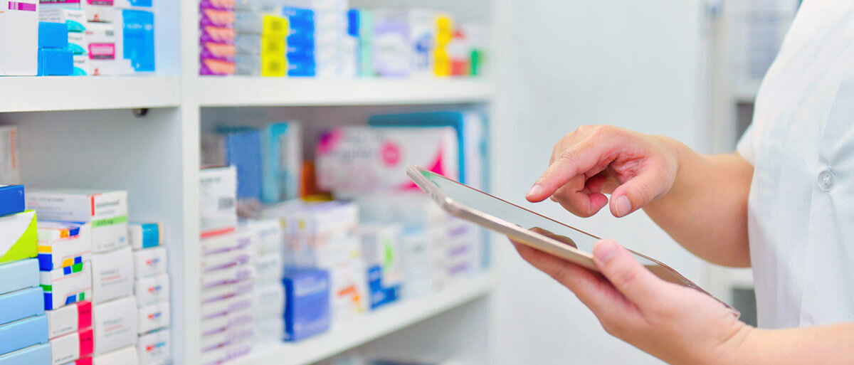 Vor einem Regal mit Fertigarzneimittelpackung steht eine Person in weißem Kittel und tippt auf einem Tablet.