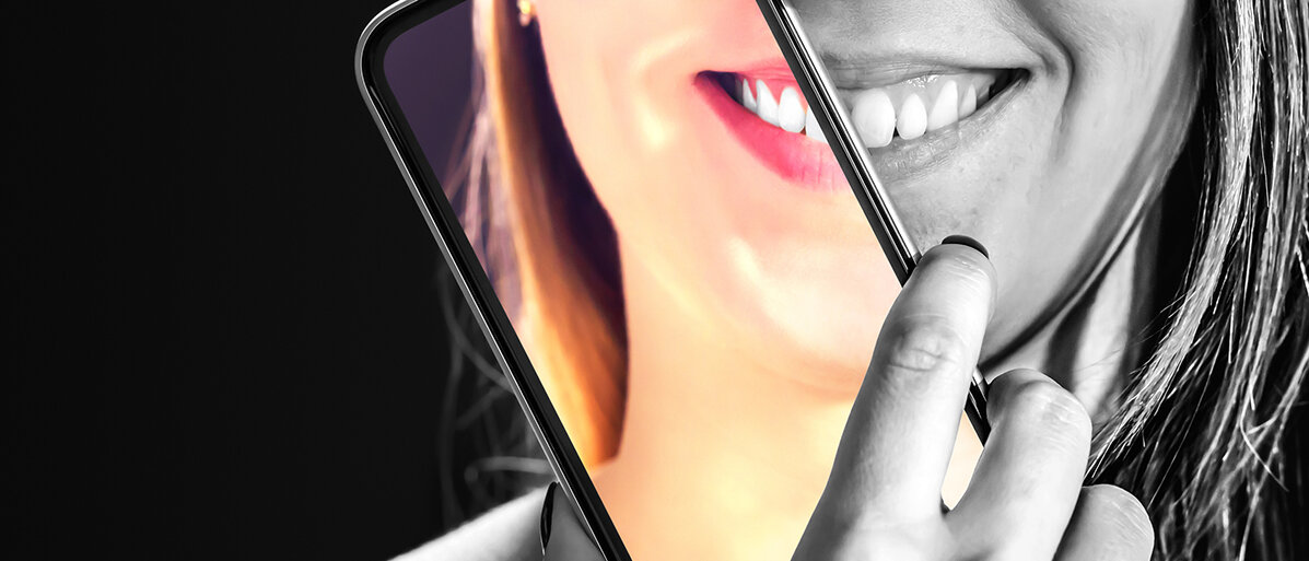 Eine lachende Frau in Schwarzweiß mit Handy vor dem Gesicht, auf diesem ist der Hautausschnitt farbig dargestellt