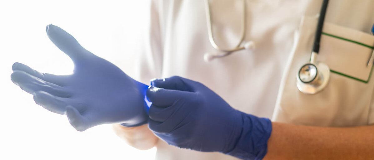 Eine Person in weißem Kittel und mit einem Stethoskop um den Hals legt blaue Nitrilhandschuhe an.