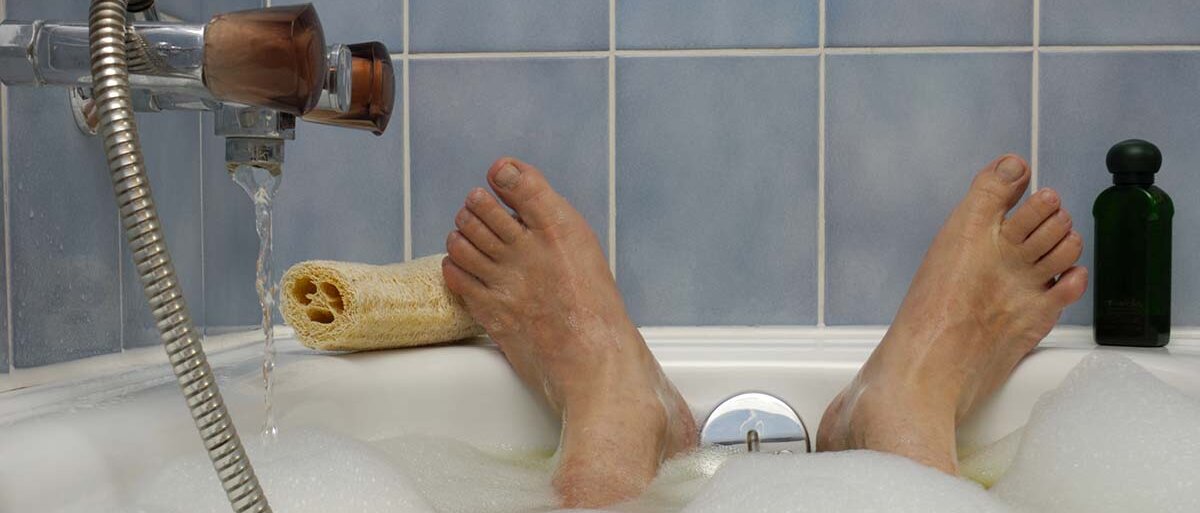 Aus Sicht einer badenden Person: Ein Haufen Schaum, der Wasserhahn der Badewanne und eine altmodisch gekachelte Wand.