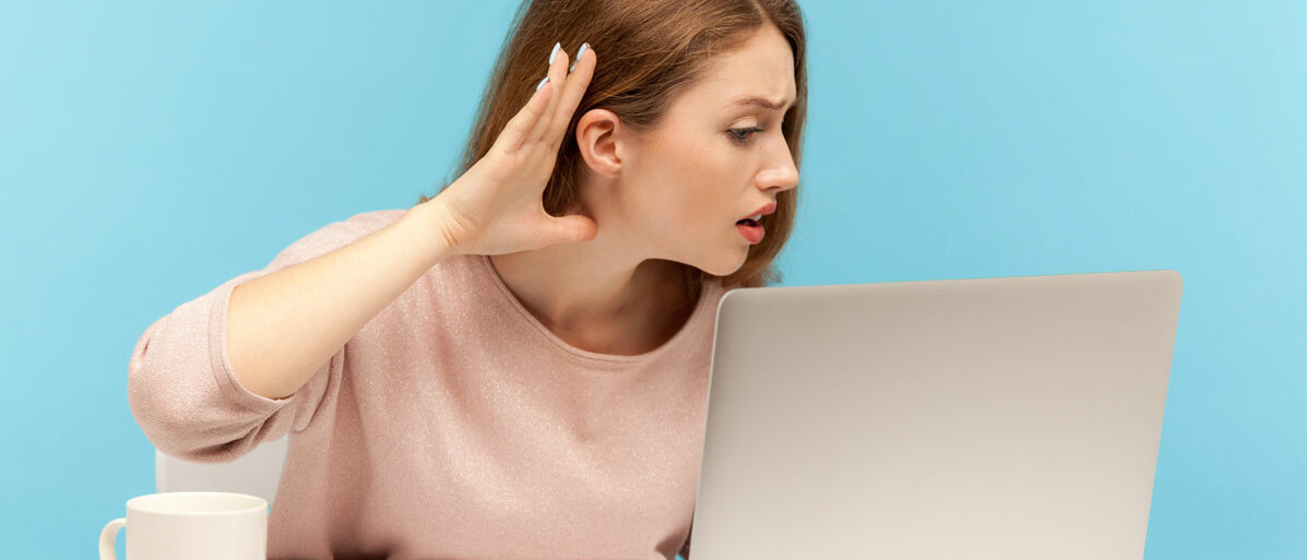 Frau sitzt vor Laptop und hält sich Hand ans Ohr weil sie schlecht hört
