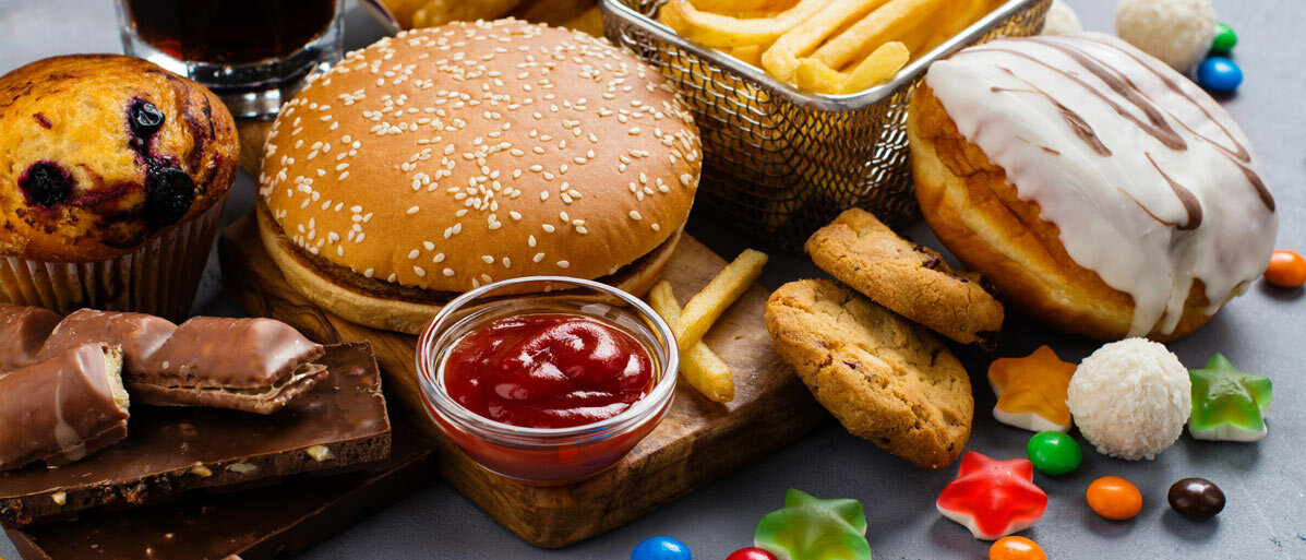 Auf einem Brett sind ein Burger, Chicken Wings, Pommes, ein Glas Cola, ein Donut, Schokoriegel, ein Muffin, Gummibärchen und Schokolinsen angerichtet.