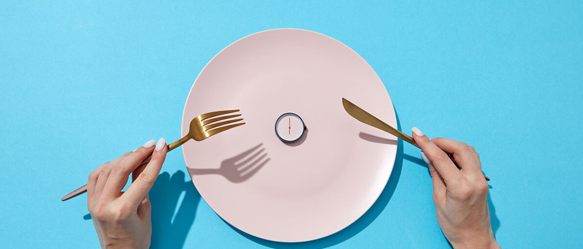 Weißer Teller mit kleiner Uhr drauf, daneben hält Frau Messer und Gabel in den Händen