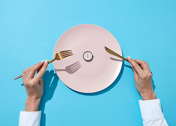 Weißer Teller mit kleiner Uhr drauf, daneben hält Frau Messer und Gabel in den Händen