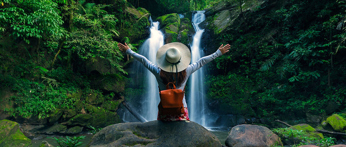 Eine Frau in Wanderkleidung sitzt vor einem Wasserfall und reckt euphorisch die Arme nach oben.