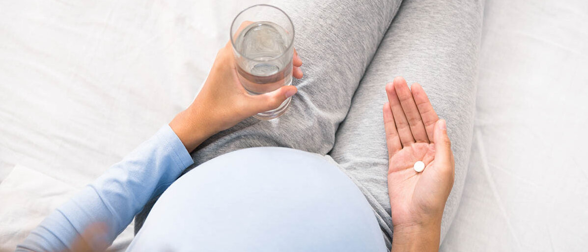 Eine schwangere Frau mit rundem Bauch hält in der einen Hand ein Glas Wasser, in der anderen eine weiße, runde Tablette.