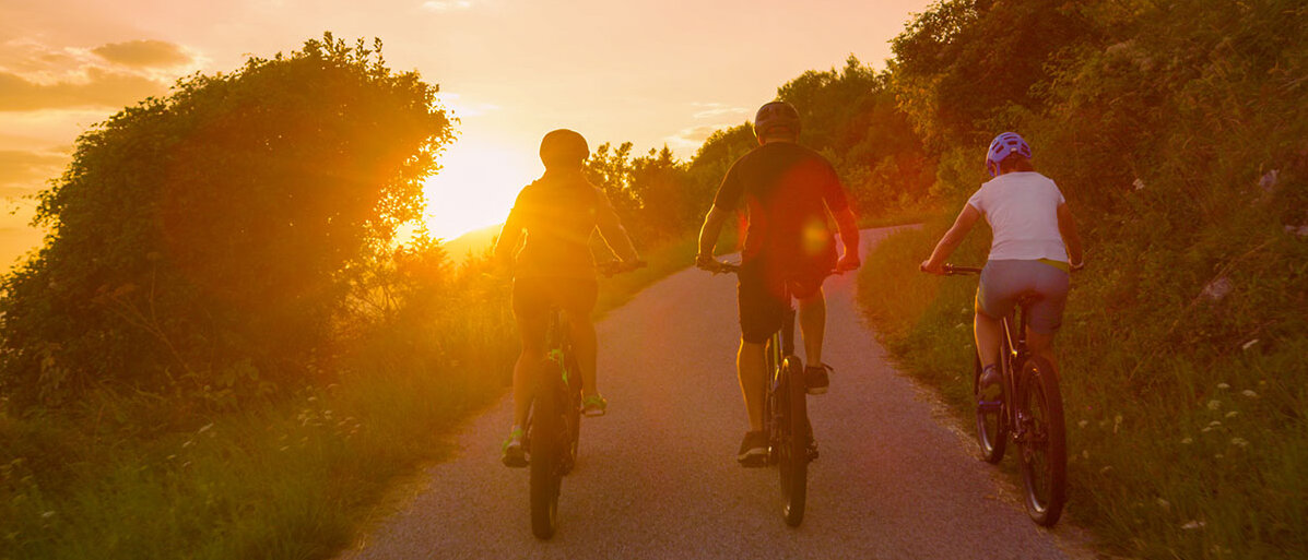 Drei Freunde fahren eine Straße bei Sonnenuntergang auf E-Bikes entlang.  