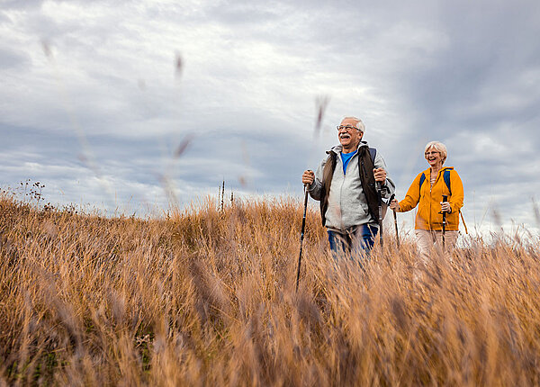 Alzheimer vorbeugen durch körperliche Aktivität älteres Paar mit Rucksäcken und Walking Stöcken beim Wandern in der Natur