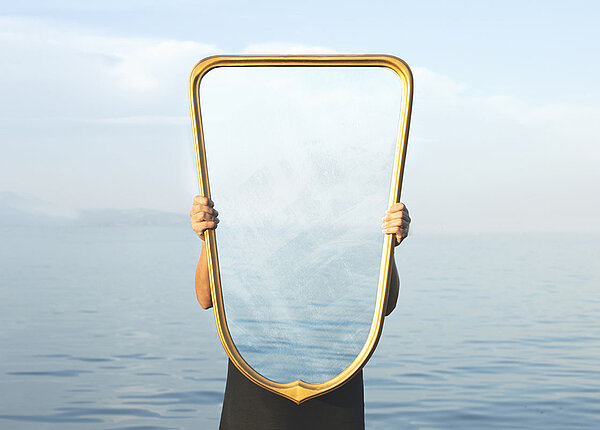 Eine Frau steht vor einem See und hält einen Spiegel in der Hand. Durch den Spiegel sehen wir aber weder die Frau noch uns selbst, sondern den See, der eigentlich hinter der Frau und hinter dem Spiegel liegt und nicht zu sehen sein dürfte.