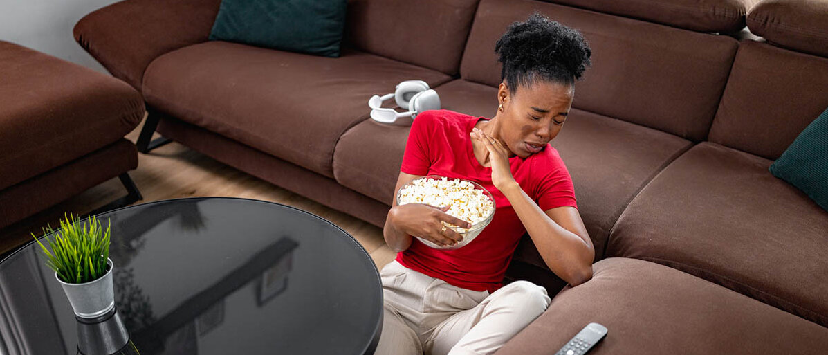 Erstickungsgefahr: Als Erste Hilfe beim Verschlucken kann der Heimlich-Griff vor dem Ersticken schützen. Eine Frau sitzt auf dem Wohnzimmerboden, hält eine Schüssel Popcorn und hat sich verschluckt.