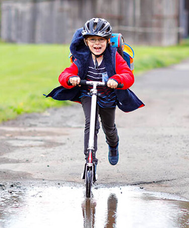 Ein Junge von ungefähr 7 Jahren fährt Roller, er trägt Helm und einen großen Schulranzen. Die Jacke flattert im Fahrtwind. Der Boden ist uneben, er steuert auf eine Pfütze zu und lacht mit weit offenem Mund.