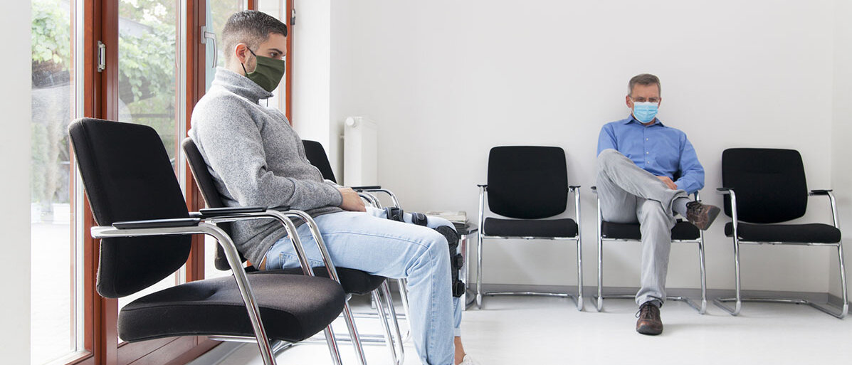 Zwei Männer sitzen mit Maske im Wartezimmer beim Arzt
