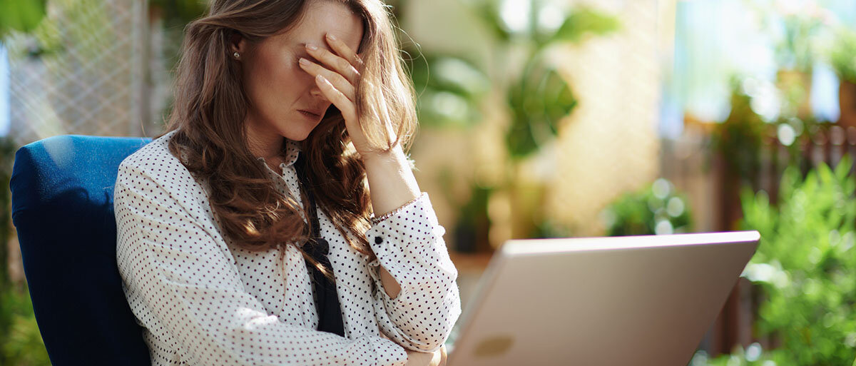 Junge gestresste Frau sitzt mit einem Laptop auf dem Schoß in ihrem Wintergarten. Sie hält sich den Kopf. 