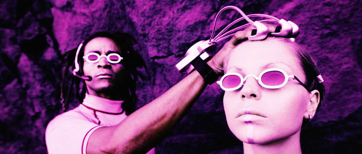Ein Mann mit Spezialhandschuh legt seine Hand auf den Kopf einer Frau. Beide tragen futuristische Brillen.